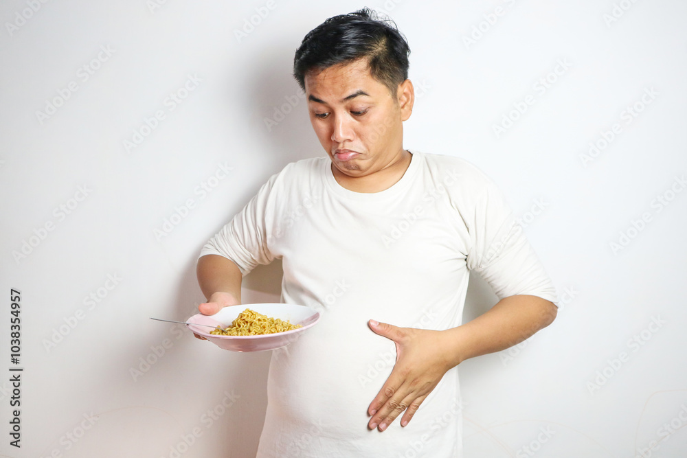 Young Asian man with noodles on plate holding stomach showing belly fat Young Asian man with noodles on plate holding stomach showing belly fat