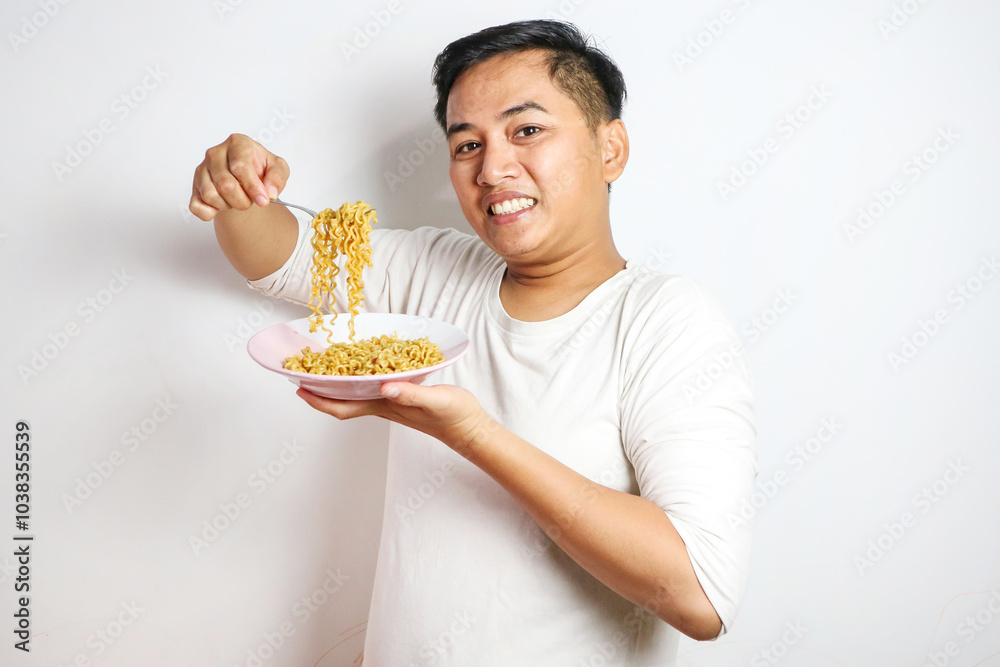 A happy, euphoric Asian young man in a tshirt eating instant fried noodles using fork isolated over a white background. stylish asian man with peek a boo hair eats instant noodle A happy, euphoric Asian young man in a tshirt eating instant fried noodles using fork isolated over a white background. stylish asian man with peek a boo hair eats instant noodle