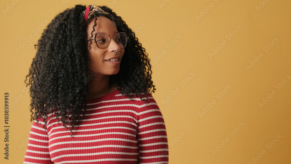 Smiling young woman with curly hair wearing glasses wearing striped sweater, looking at the free space for advertising, on yellow background in the studio Smiling young woman with curly hair wearing glasses wearing striped sweater, looking at the free space for advertising, on yellow background in the studio