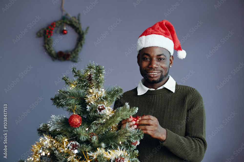Black person standing next to Christmas tree, smiling and holding ornament while wearing Santa hat while festive wreath hanging on wall in background Black person standing next to Christmas tree, smiling and holding ornament while wearing Santa hat while festive wreath hanging on wall in background