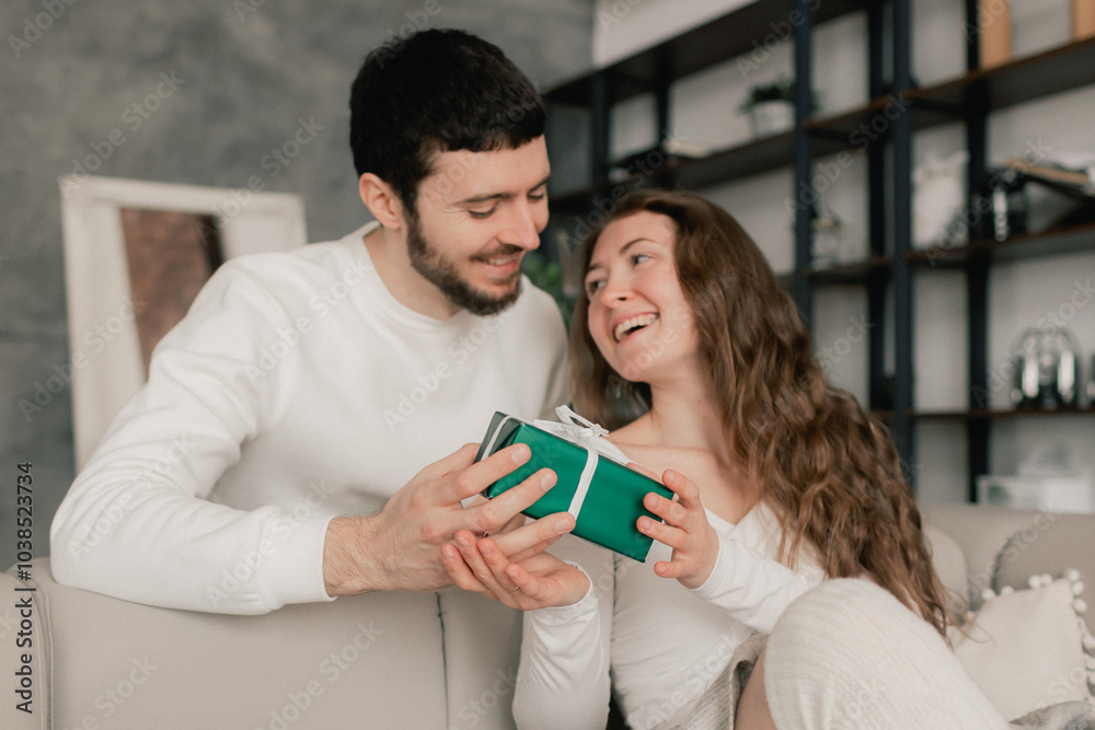 Young woman hugs her beloved husband giving him a gift on this special day. Concept of gifts for birthdays, Christmas, Valentine's Day and Men's Day. Modern living room interior. . Part of a series Young woman hugs her beloved husband giving him a gift on this special day. Concept of gifts for birthdays, Christmas, Valentine's Day and Men's Day. Modern living room interior. . Part of a series