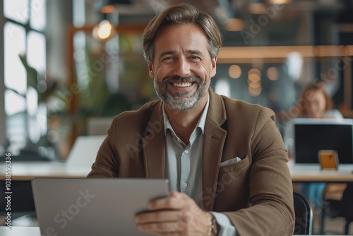 Happy businessman smiling with tablet, phone, and laptop inside the modern office.