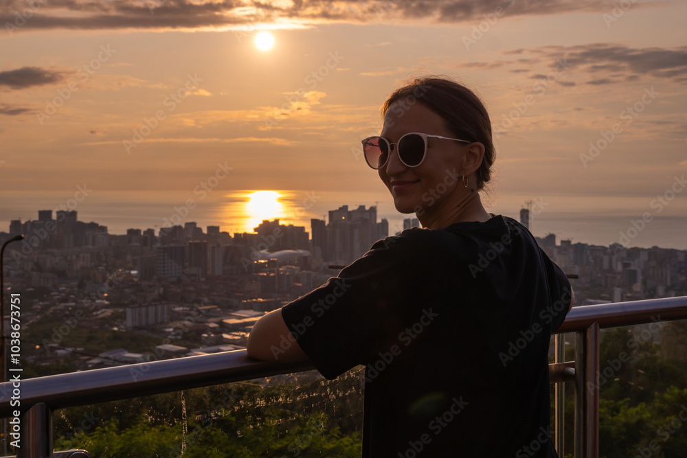 Fototapeta premium Young woman is leaning on a railing, enjoying a beautiful sunset over the cityscape. Sun is reflecting on the sea creating a romantic atmosphere