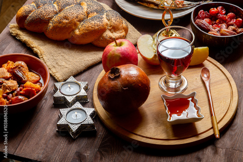 Challah, honey, apple and pomegranate and wine and chelnt on the table with menorah and burning candles in honor of Rosh Hashanah