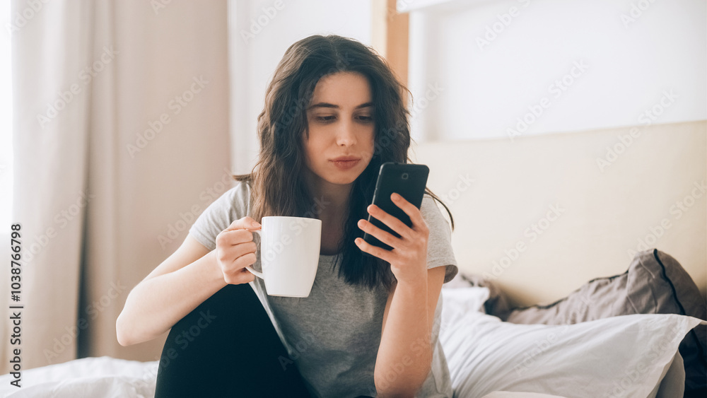 Mobile communication. Internet browsing. Gadget rest. Concentrated woman reading social media on phone drinking coffee on bed light bedroom interior. Mobile communication. Internet browsing. Gadget rest. Concentrated woman reading social media on phone drinking coffee on bed light bedroom interior.