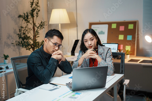 Two young businesspeople work late in the office, visibly worried, focusing intently on laptops, trying to find solutions