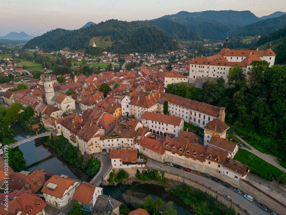 Beautiful Skofja Loka medieval town with church and the castle at sunset. Aerial view of riverfront with historic houses and the bridges over Sora river. Hilly landscape towards Smarna Gora.