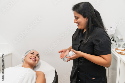 Beautician showing skincare product to woman relaxing on bed