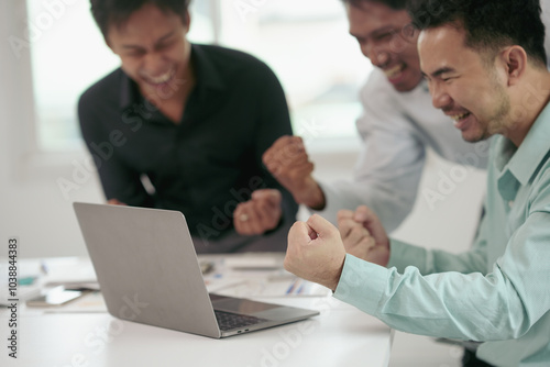 Diverse group of colleagues celebrating a major success in a vibrant office setting, gathered around a laptop, exchanging high fives and fist pumps to mark their achievement