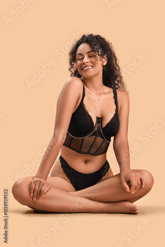 Beautiful young happy African-American woman in stylish lace lingerie sitting on beige background