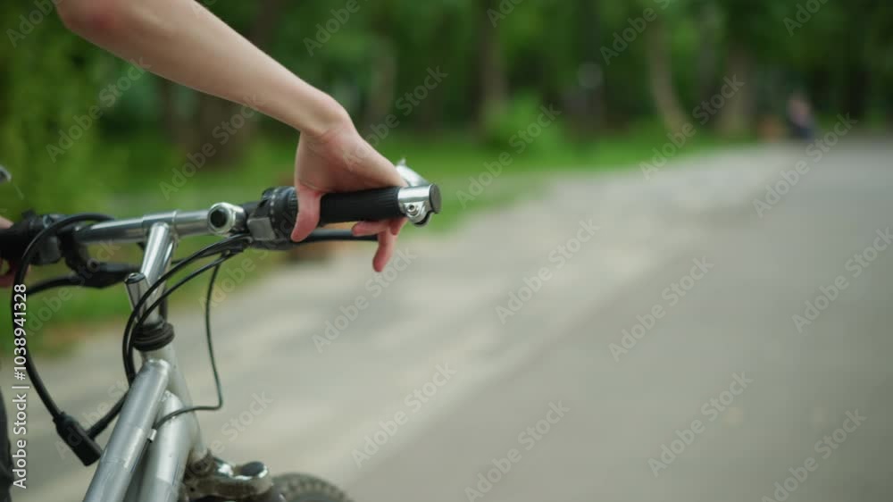 Partial view of someone in black top walking closely with his bicycle along a calm, peaceful pathway, with focus on his hand holding the handlebar of bicycle, the road is surrounded by greenery