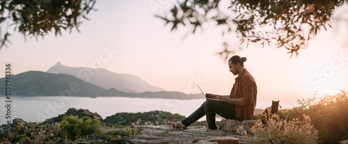 A Caucasian man is working with a laptop in a garden on a mountain overlooking the sea and sunset.