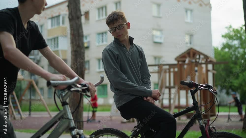 Two boys are sitting on their bikes, one boy rests while holding his handlebars and looking towards the other boy, in the background, children are riding a scooter near a playground Two boys are sitting on their bikes, one boy rests while holding his handlebars and looking towards the other boy, in the background, children are riding a scooter near a playground