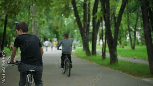 Wallpaper Mural Back view of two friends riding bicycles along a peaceful park path, with the one in the grey top looking back at his friend, people are visible walking ahead, and the setting is surrounded by trees Torontodigital.ca