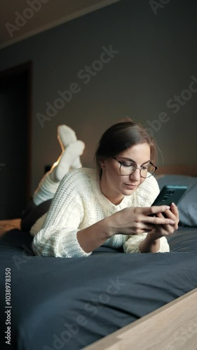 Attractive Caucasian girl spends her day off on the bed with a mobile phone. Lifestyle, day off. Vertical frame