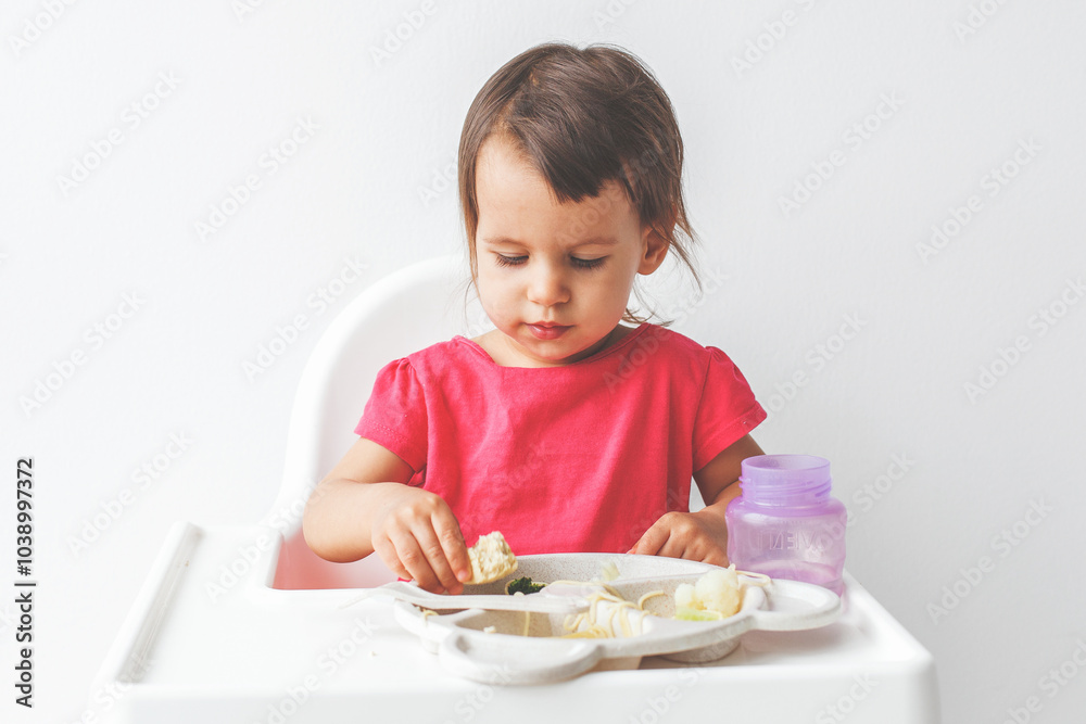 girl child in a bright pink t-shirt sitting in a high chair on a white background and having lunch, baby food concept girl child in a bright pink t-shirt sitting in a high chair on a white background and having lunch, baby food concept