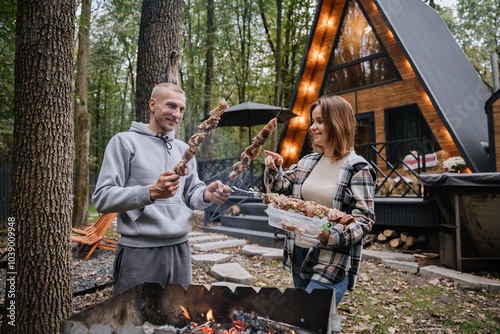 Couple preparing skewers for barbecue over open fire at cabin, enjoying autumn grilling together