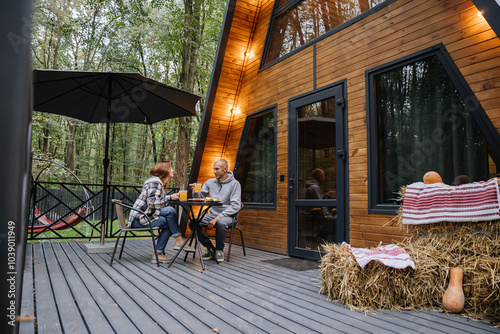 Couple enjoying meal on wooden terrace of A-frame cabin in the forest, creating a cozy autumn vibe
