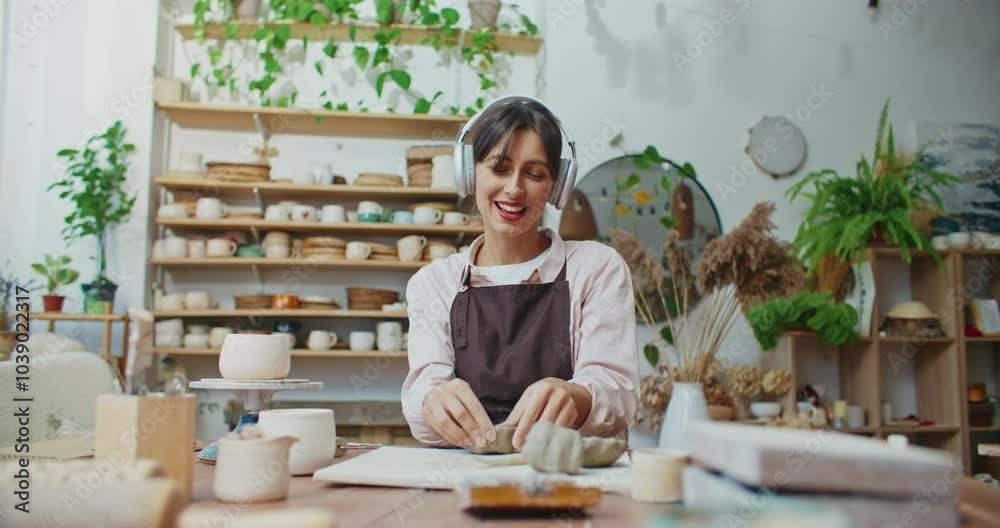 Positive Caucasian girl sitting at desk and carefully pressing with fingers piece of clay. Finishing creating small bowl. Listening to music while working in studio as potter. Wearing headphones. Positive Caucasian girl sitting at desk and carefully pressing with fingers piece of clay. Finishing creating small bowl. Listening to music while working in studio as potter. Wearing headphones.
