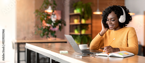 Happy black girl in wireless headphones studying online, using laptop and taking notes, cafe interior, copy space photo