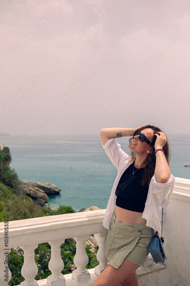 Beautiful girl posing on a balcony overlooking the sea. Girl in sunglasses and a white blouse enjoying a seaside vacation in Spain. Vacation at sea in the velvet season Beautiful girl posing on a balcony overlooking the sea. Girl in sunglasses and a white blouse enjoying a seaside vacation in Spain. Vacation at sea in the velvet season