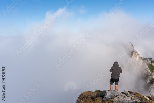 Hiker standing on a misty mountain ridge, gazing into the clouds with a clear view of the sharp peak ahead on a bright day. Concept of mountain exploration, adventure travel, connection with nature. 