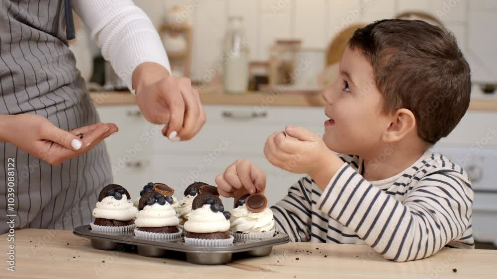 A cheerful boy smiles as he decorates cupcakes with toppings alongside an adult in a warmly lit kitchen. A cheerful boy smiles as he decorates cupcakes with toppings alongside an adult in a warmly lit kitchen.