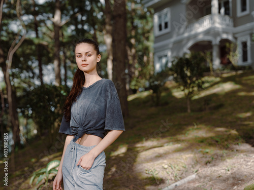 Young woman standing outdoors in a relaxed pose, wearing a loose grey shirt and sweatpants, surrounded by lush greenery and a house in the background