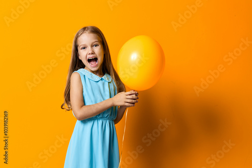 Joyful young girl in blue dress holding a bright yellow balloon against a vibrant orange backdrop photo