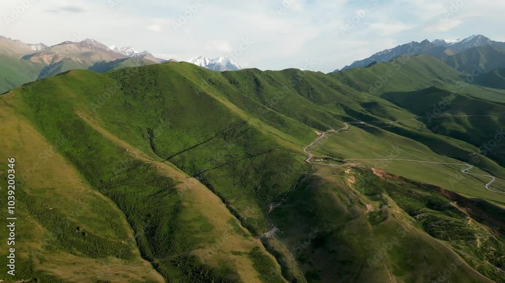 Winding road through mountain pass, difficult route in countryside. Nature of Kyrgyzstan: green hills in foreground and snow-capped peaks in distance. Summer mountains natural landscape aerial view Winding road through mountain pass, difficult route in countryside. Nature of Kyrgyzstan: green hills in foreground and snow-capped peaks in distance. Summer mountains natural landscape aerial view