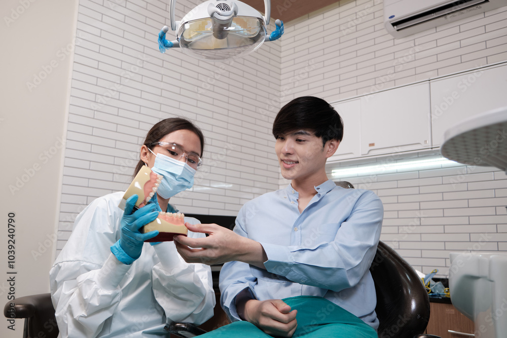 Asian female dentist explaining orthodontic care and whitening to young male patient with teeth model in dental clinic, well-being hygiene, and professional healthcare work in doctor office hospital. Asian female dentist explaining orthodontic care and whitening to young male patient with teeth model in dental clinic, well-being hygiene, and professional healthcare work in doctor office hospital.