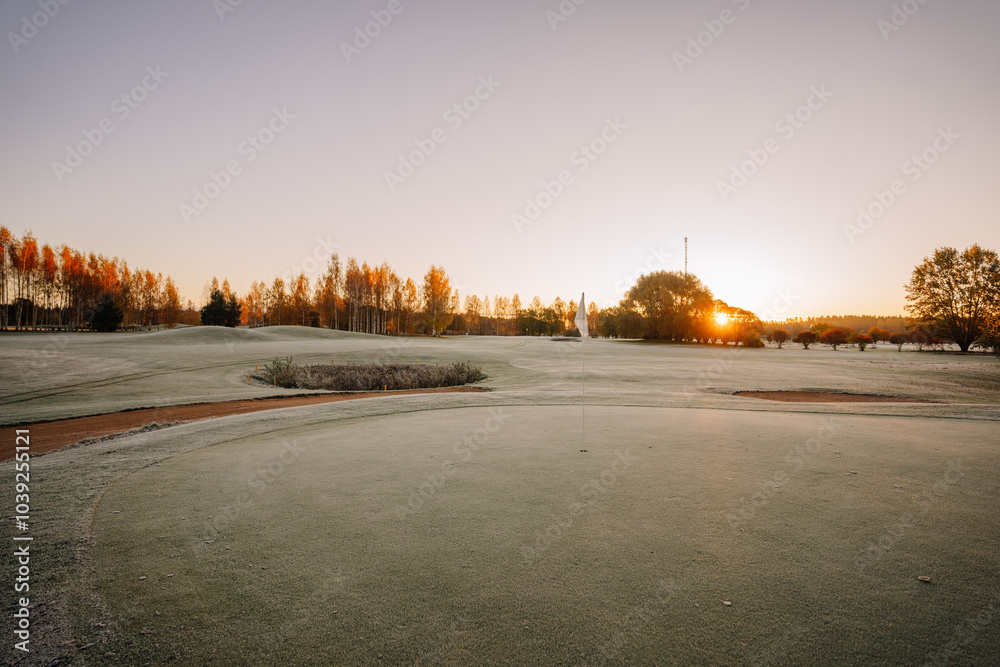 A frosted golf course at sunrise, with a flag on the green and trees in the distance illuminated by the morning sun..