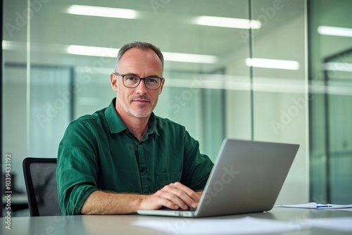  Middle-aged man with glasses working on a laptop at an office desk, blurred glass wall background, wide-angle professional photograph.