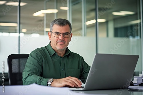  Middle-aged man with glasses working on a laptop at an office desk, blurred glass wall background, wide-angle professional photograph.