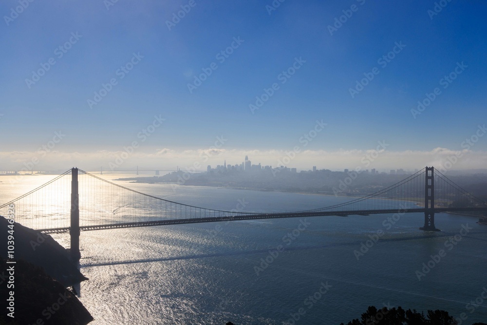 Obraz premium Bright sun reflects on bay behind tower of Golden Gate Bridge with city in distance