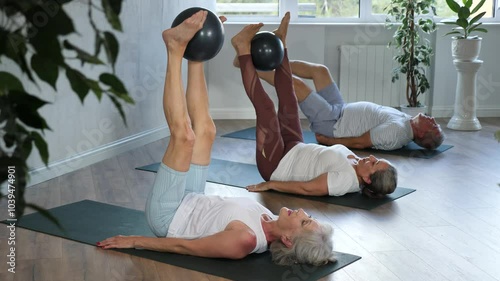 Senior woman in company of elderly friends and daughter perform Pilates exercises with softball. Visitior yoga class lying on back, raised legs perpendicular to floor and clamped softball in feet photo