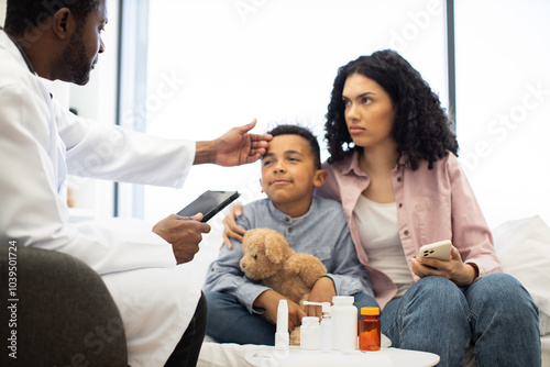 Male doctor examining young African American boy sitting with mother. Child holding teddy bear, medical supplies on table. Mother appearing concerned holding smartphone.