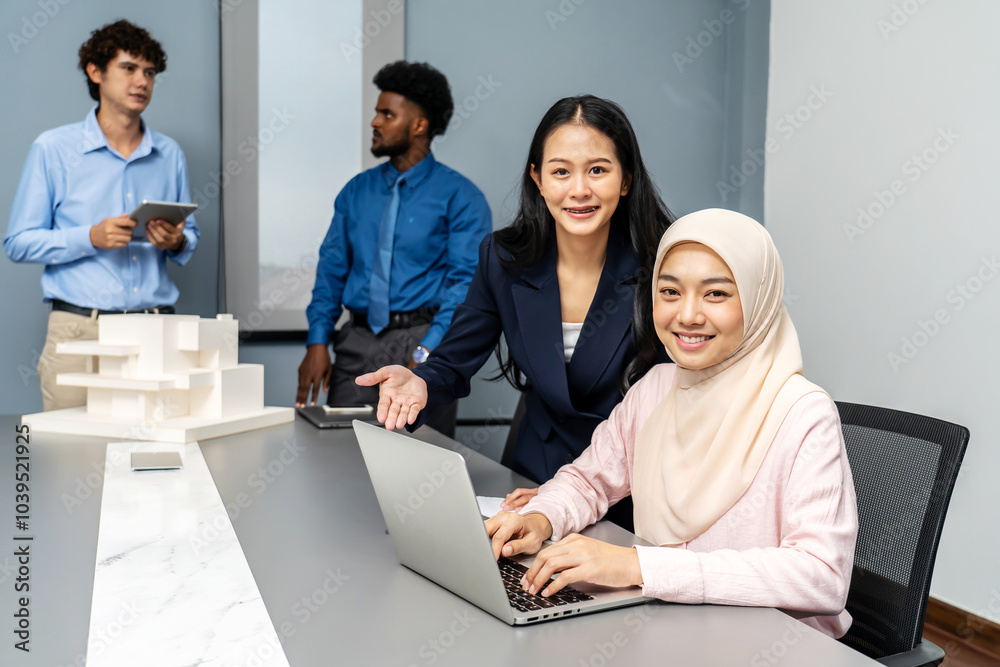 Smiling asian and muslim women working together on a laptop, With a background as male colleagues discussing house model in office. They checking data in computer at meeting room together, look camera Smiling asian and muslim women working together on a laptop, With a background as male colleagues discussing house model in office. They checking data in computer at meeting room together, look camera