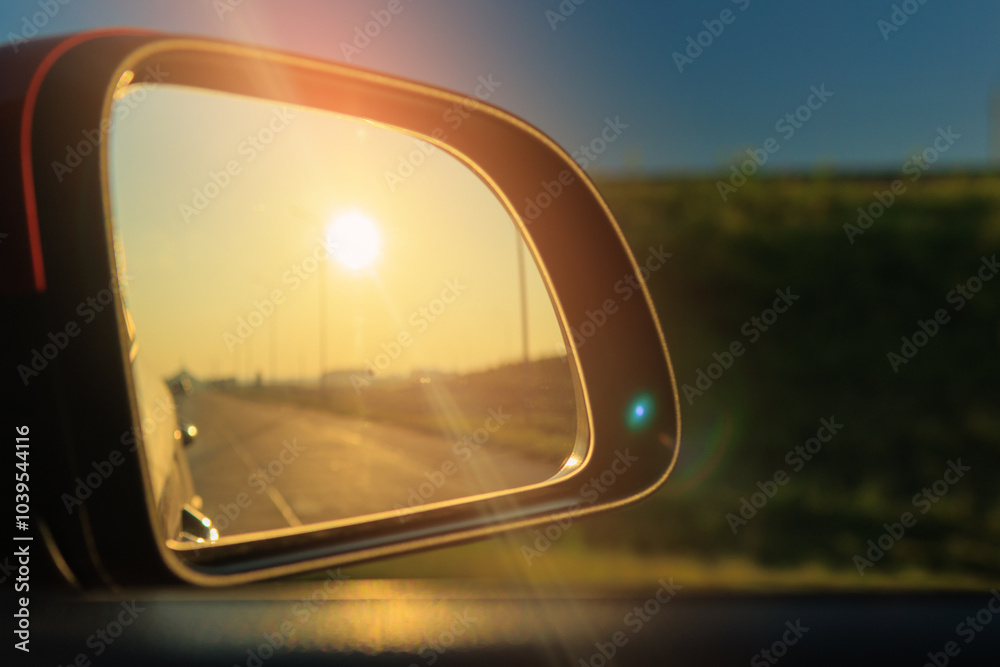 The sun is reflected in the mirror, evening road. View in the side rear view mirror of a red car driving along the highway and street The sun is reflected in the mirror, evening road. View in the side rear view mirror of a red car driving along the highway and street