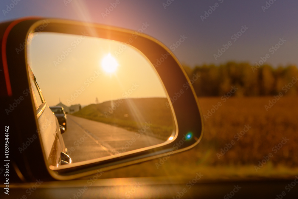The sun is reflected in the mirror, evening road. View in the side rear view mirror of a red car driving along the highway and street