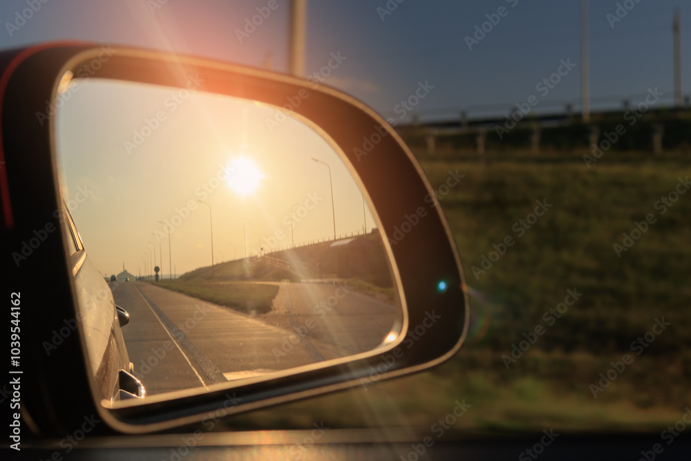 The sun is reflected in the mirror, evening road. View in the side rear view mirror of a red car driving along the highway and street The sun is reflected in the mirror, evening road. View in the side rear view mirror of a red car driving along the highway and street