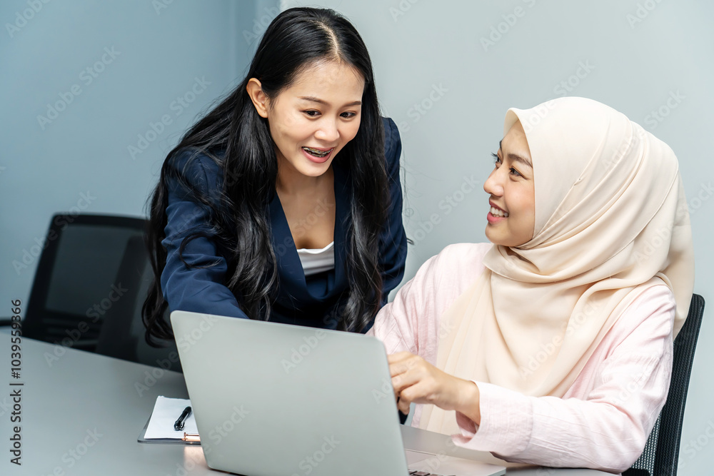 Smiling asian and muslim women working together on a laptop, They checking data in computer at meeting room together, using a laptop for a productive business meeting in a corporate workplace. Smiling asian and muslim women working together on a laptop, They checking data in computer at meeting room together, using a laptop for a productive business meeting in a corporate workplace.