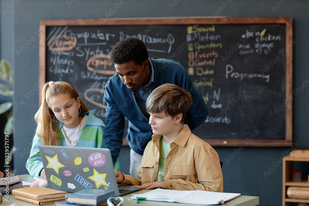 Portrait of African American male teacher helping two teenage kids using laptop together during lesson in school classroom Portrait of African American male teacher helping two teenage kids using laptop together during lesson in school classroom