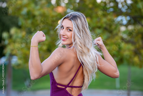 Smiling blonde athlete woman flexing her biceps after training outdoor in a park photo