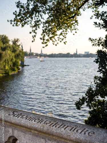 Alster Lake in Hamburg during evening with distant sailboats, framed by greenery filtering sunlight through the trees and a stone bridge railing featuring the sharp Schwanenwikbrücke inscription. photo