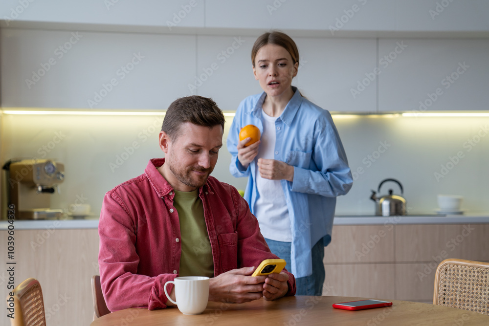 Woman secretly reads husband messages over shoulder while he chats on kitchen. Suspicion, trust issues, relationship doubts, emotional distance, betrayal fears, jealousy, invasion of privacy Woman secretly reads husband messages over shoulder while he chats on kitchen. Suspicion, trust issues, relationship doubts, emotional distance, betrayal fears, jealousy, invasion of privacy