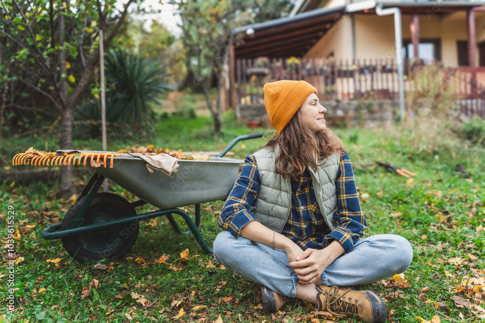 Young happy woman collecting autumn leaves in the backyard of a country house using a rake and cart