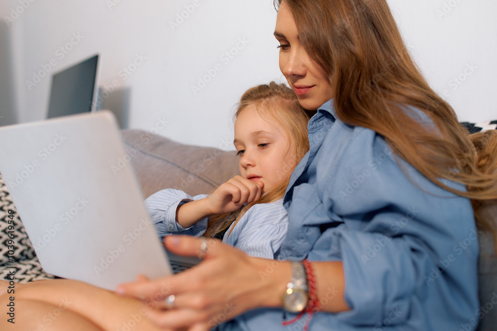 Young mother and daughter watching video on laptop while sitting on sofa Young mother and daughter watching video on laptop while sitting on sofa