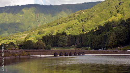 The photo shows a bridge over a huge blue-green lake, surrounded by the green slopes of the Portuguese Azores. The photo is taken from a distance, showing nature and a blue sky. photo