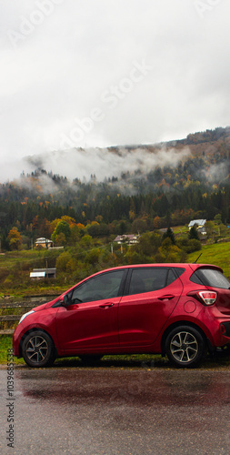 Red car on the road in the mountains on rainy day. Autumn travel by car. Hatchback car in mountain village with clouds. Road journey in picturesque Carpathian mountains. Car on wet road in countryside